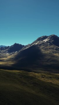 A wide view shows mountains and a valley filled with grass under a clear blue sky. The sun begins to rise, casting long shadows across the landscape. The scene captures natures beauty.