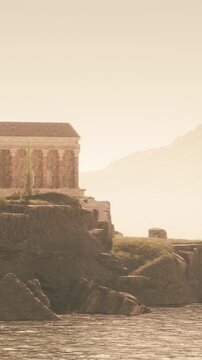 A stone temple sits on a rocky cliff by the sea. The sun sets behind distant hills, casting a soft light over the landscape. Water laps at the shore, and trees grow near the temple.