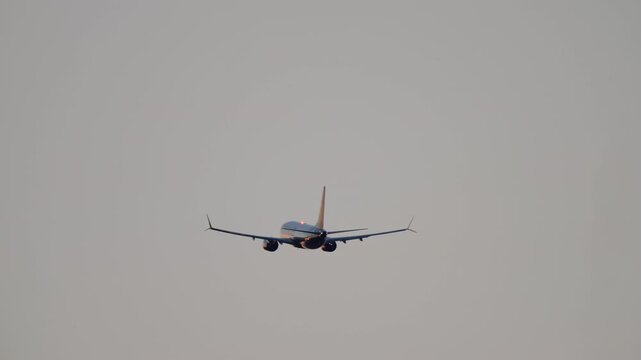 Passenger jet climbing after takeoff, rear view against grey overcast sky, swept wings with winglets and twin engines, airliner departing from the airport in hazy evening light