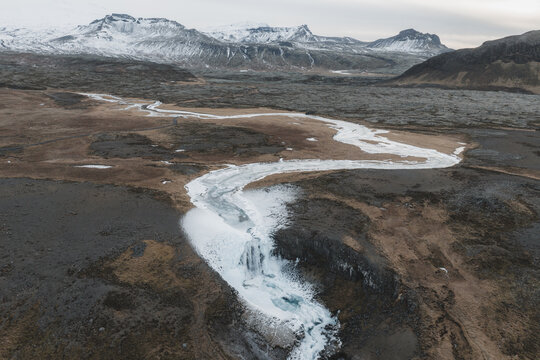 Aerial view of a frozen waterfall cascading through a rugged volcanic landscape with snow-capped mountains in the background, Iceland.