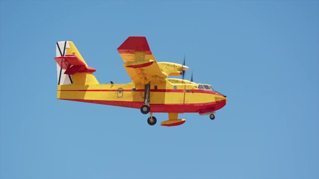 Yellow and red amphibious firefighting plane in flight, side view with landing gear extended, twin turboprop water bomber against clear blue sky, slow motion