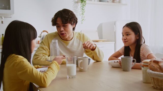 Smiling Asian Children Enjoying Breakfast Together With Brother At Cozy Kitchen Table, Eating Cereal And Drinking Milk, Creating Joyful Moments In Family Setting. Family Lifestyle Concept.