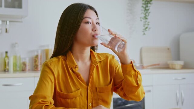 Focused Young Asian Woman Taking A Sip Of Water From A Glass, Sitting At Kitchen Table In Modern Kitchen. Emphasizing Importance Of Hydration In Daily Routine, Healthy Lifestyle Concept.
