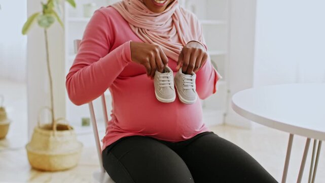 Smiling African American Woman In Pink Top Holding Baby Shoes, Sitting On Chair At Home, Anticipating Arrival Of Newborn, Feeling Excited And Joyful In Cozy Living Space. Expecting Concept.