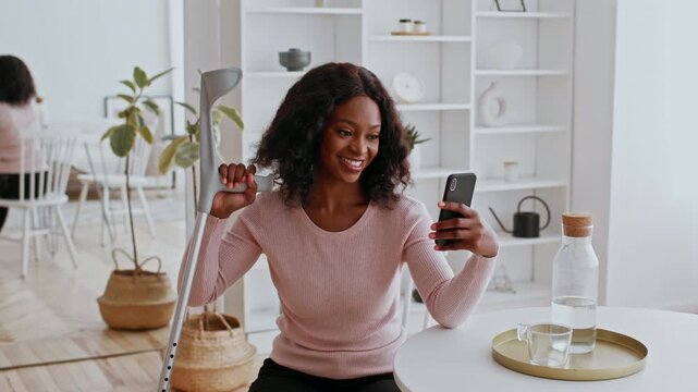 Happy Black Woman Smiling While Using Smartphone At Home, Sitting At Modern Table With Indoor Plants. Engaging Interaction, Indoor Lifestyle Setting, Cheerful Mood Emphasized.