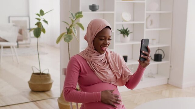 Smiling Young Black Woman Holding Smartphone While Touching Her Pregnant Belly At Home, Showcasing Joy And Anticipation In A Modern Living Room Setting. Maternity Concept.