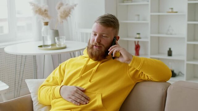 Happy Bearded Man Talking On Phone At Home, Sitting On Couch In Modern Living Room, Relaxed And Comfortable, Engaging In Casual Conversation, Home Lifestyle Concept.