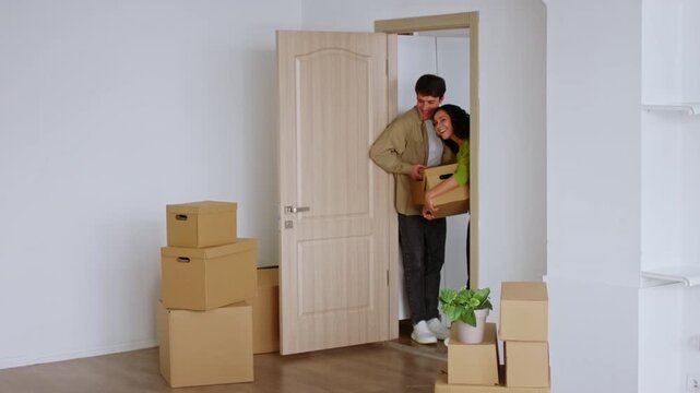 Smiling Young Couple, One Man And One Woman, Carrying Boxes While Moving Into Their New Apartment, Joyfully Entering A Minimalist Space Filled With Moving Boxes. Home Concept.