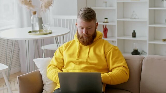 Focused Caucasian Male In Yellow Hoodie Working On Laptop At Home, Sitting Comfortably In Living Room Setting. Engaged In Professional Work, Emphasizing Remote Business Concept.