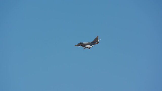 Fighter jet flying in vast clear blue sky, small side profile of delta wing combat aircraft with canards, landing gear lowered on approach, slow motion
