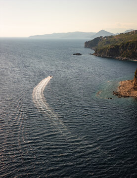 Aerial view of a small boat leaving a white wake on the deep blue Adriatic Sea near the rugged coastline and cliffs of Petrovac, Budva Municipality, Montenegro.