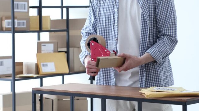 Man using dispenser with adhesive tape to seal cardboard box before sending at table in office, closeup