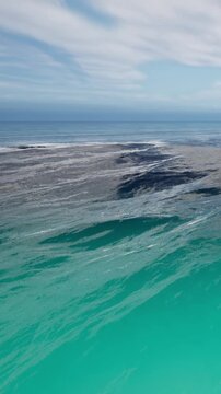 Waves crash against rocks close to the shore under a clear sky. The water shows different shades of blue as sunlight reflects on the ocean surface.