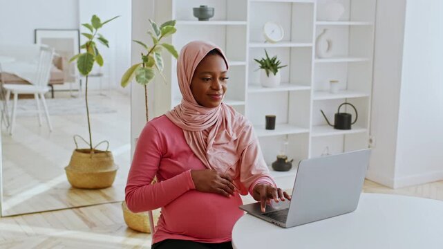 Focused Pregnant African American Woman Using Laptop While Sitting At Home. Comfortable Atmosphere With Indoor Plants And Modern Decor, Engaging In Work Or Study Activity. Healthcare Concept.