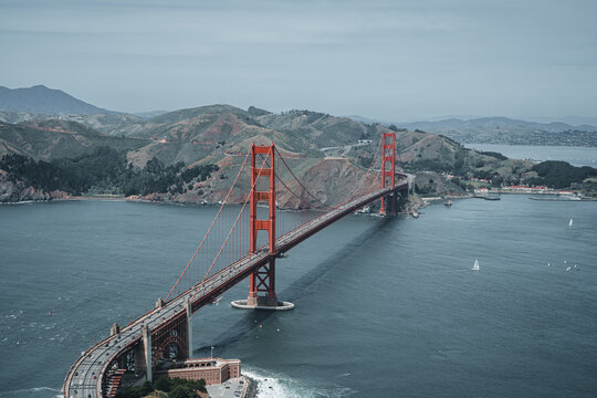 Aerial view of the Golden Gate Bridge spanning the bay with rolling hills in the background under a cloudy sky in San Francisco, California, United States.