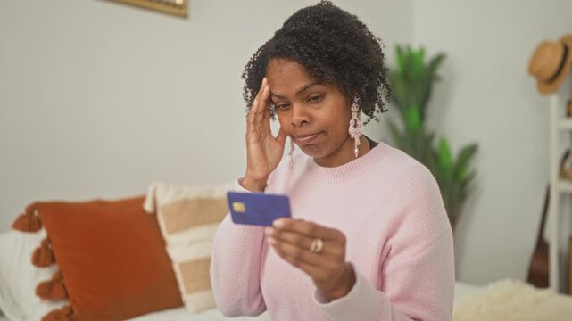 Young african american woman sitting on bed holding creditcard and touching temple while inspecting chip and card details in building; budgeting worry.