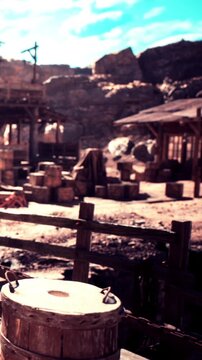 In a dusty old western town, wooden structures and barrels fill the scene. The sun shines brightly, highlighting the rugged terrain and buildings that create a vintage atmosphere.