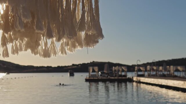 Close-up shot of white macrame fringe swaying gently with blurred luxury beach club and calm bay in golden background light, Aegean coast Turkey.