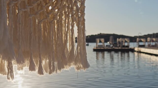 Close-up shot of white macrame fringe swaying gently with blurred luxury beach club and calm bay in golden background light, Aegean coast Turkey.