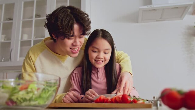 Smiling Asian Young Man And Girl Actively Preparing Food Together In Modern Kitchen, Enjoying Each Other's Company While Cooking Healthy Meal. Family Bonding Experience In Cozy Environment.