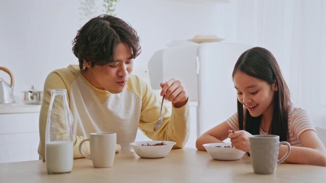 Happy Young Asian Boy And Girl Eating Breakfast Together At Home, Sharing A Bowl Of Cereal And Milk At Kitchen Table, Enjoying Quality Time While Developing Healthy Eating Habits.