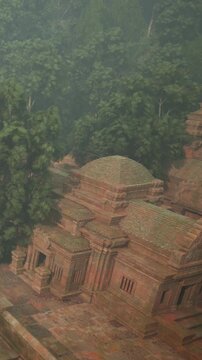 An ancient temple stands among thick trees in a forest. The temple is made of red stone and features a distinct architectural style. The area is quiet and natural, filled with greenery.
