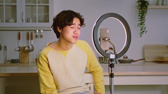 Focused Male Asian Creator Recording Video With Smartphone On Ring Light Stand In Modern Kitchen, Capturing Content And Engaging With Audience In Digital Space. Professional Content Creation Concept.