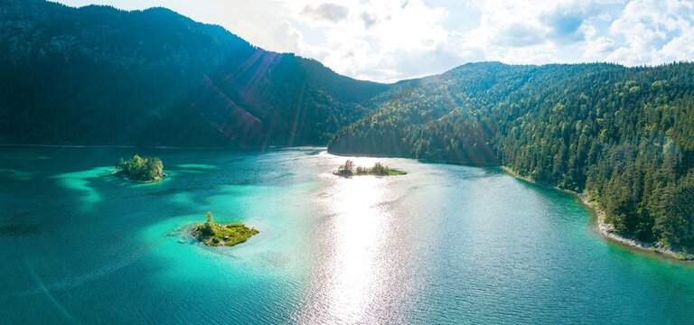 Aerial view of Eibsee with its turquoise waters and small forested islands surrounded by dense pine forests and mountain slopes under bright sunlight in Grainau, Bavaria, Germany.