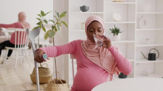 Focused Pregnant Black Woman Using Crutch And Drinking Water At Home, Sitting At Kitchen Table In Modern Environment, Prioritizing Health And Wellness During Pregnancy.