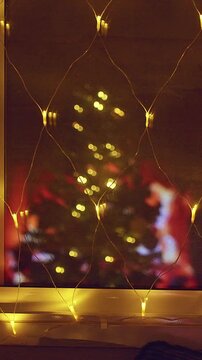 Vertical video. Golden Fairy Lights on a Window With Reflections of a Christmas Tree and Fireplace, Cozy Holiday Interior, Warm Winter Evening and Festive Spirit
