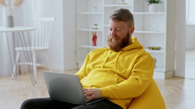 Smiling Young Caucasian Man Sitting Comfortably In Cozy Setting, Using Laptop For Work, Enjoying Remote Freelancing In Modern Home Atmosphere. Focused On Screen, Relaxed Mood.