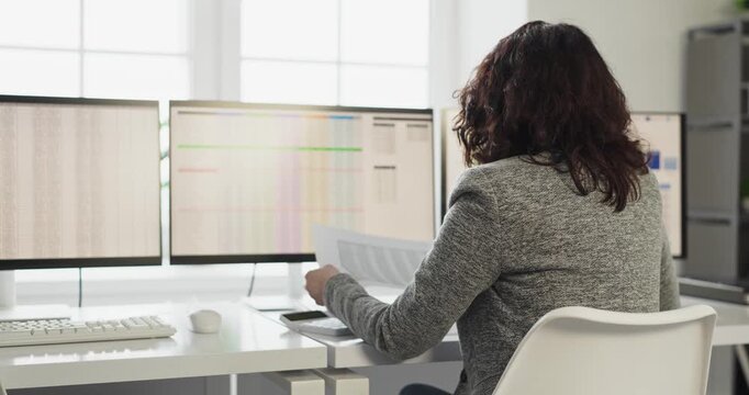 Woman analyst working office monitors spreadsheets. Rear view of professional reviewing finance data on multi displays, holding paperwork at clean desk. Clear focus on audit and data reports.
