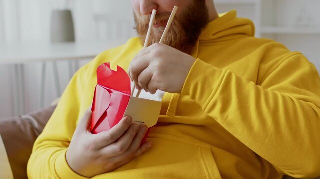 Focused Man With Beard Eating Asian Noodles With Chopsticks While Sitting In Comfortable Setting At Home, Enjoying A Meal. Weight Loss Concept.