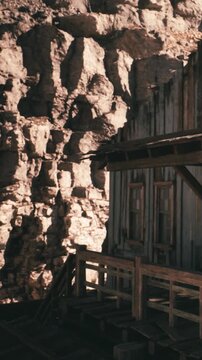 A wooden structure stands next to a large rock formation. Sunlight brightens the area. The building shows signs of age. The setting is rustic and natural, with a clear sky above.