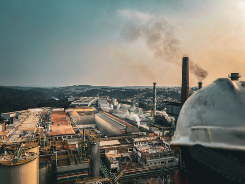 Industrial engineer or safety supervisor wearing a hard hat overlooking a large processing plant with smokestacks and city background.
