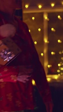 Vertical video. Cheerful Woman Dancing With a Wrapped Christmas Gift in Front of a Decorated Tree and Warm Fairy Lights, Cozy Holiday Home, Festive Spirit and Family Joy