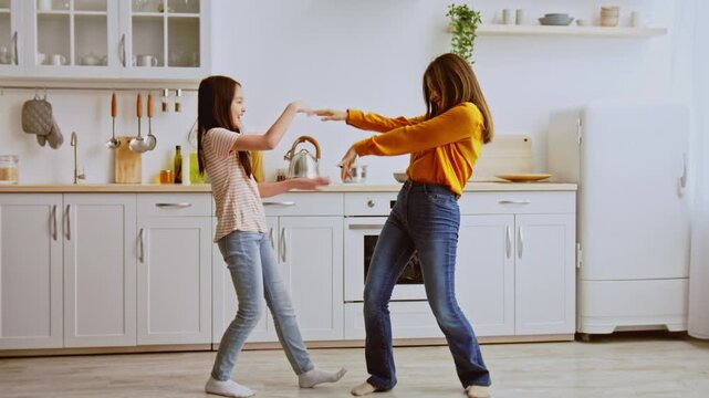 Joyful Happy Young Asian Girl And Woman Dancing Together In Bright Kitchen, Enjoying Fun Time While Listening To Music, Creating Beautiful Memories With Family In Cozy Home Setting.