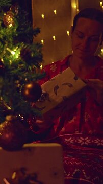 Vertical video. Happy Woman Unwrapping a Christmas Gift Beside a Decorated Tree With Warm Golden Fairy Lights, Cozy Holiday Home, Festive Celebration and Family Tradition