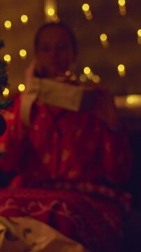 Vertical video. Young Woman Reaching With Joy to Receive a Wrapped Christmas Gift Beside a Decorated Tree With Warm Fairy Lights, Cozy Holiday Home, Giving, Love and Family Celebration