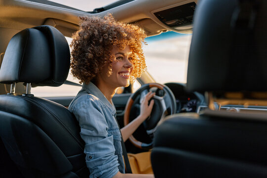 Smiling woman with curly hair sits behind the wheel of a car, wearing a denim shirt. Warm interior lighting emphasizes daily lifestyle, confidence, and self care on the road.