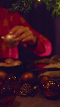 Vertical video. Christmas Ornaments Spread Across a Table as a Woman Prepares Decorations for a Festive Tree, Cozy Holiday Atmosphere and Warm Fireplace