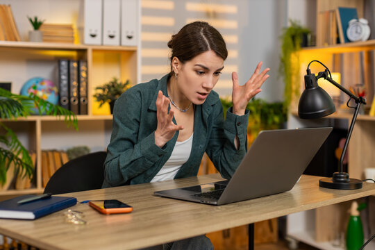 Young woman on home table looks at laptop with angry face after online work error and hard problem in living room. Adult girl on home table feels stress frustration and anger during remote job task.