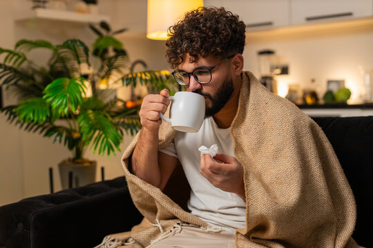 Arabian man at home feels sore throat holds mug of hot tea with medicine takes slow sip uses tissue. Egyptian guy on sofa searches doctor on smartphone rubs neck coughs rests under blanket sniff fever