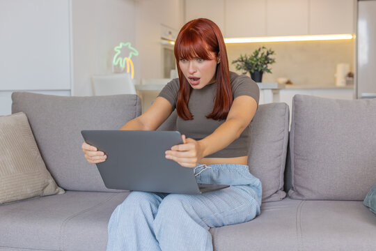 Young woman at home hits laptop freeze virus glitch causes crash feels despair stress and anger. Redhead girl on sofa tries restart and scan yet system fails tasks stop rush fix wait frustration loud.