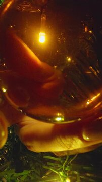 Vertical video. Close Up View From Inside a Christmas Tree as Female Hands Carefully Hang a Golden Holiday Bauble Among Glowing Evergreen Branches, Christmas Decoration