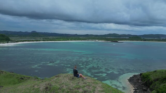 Woman Sitting Cliff Overlooking Turquoise Bay, Panoramic Aerial Viewpoint Showing Coral Reef Patterns, Distant Green Hills And Crescent Beach,Bukit Merese, Lombok, Tanjung Ann
