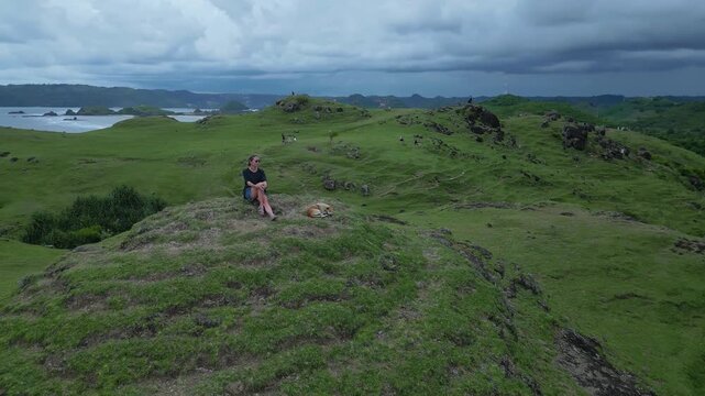 Caucasian Woman Contemplating Stormy Bay, Sits With Dog On Ridge Under Brooding Clouds, Dramatic Ocean Tones And Distant Islands Create Reflective Mood And Wanderlust Companionship,Lombok