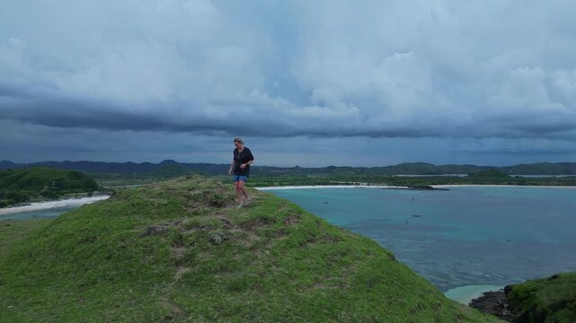 Woman Surveying Wide Turquoise Bay From Hilltop With Expansive Lagoon And Distant Shoreline Solitary Figure On Crest, Curving Paths In Grass, Calm Sea Tones, Cinematic Aerial Vista, Lombok