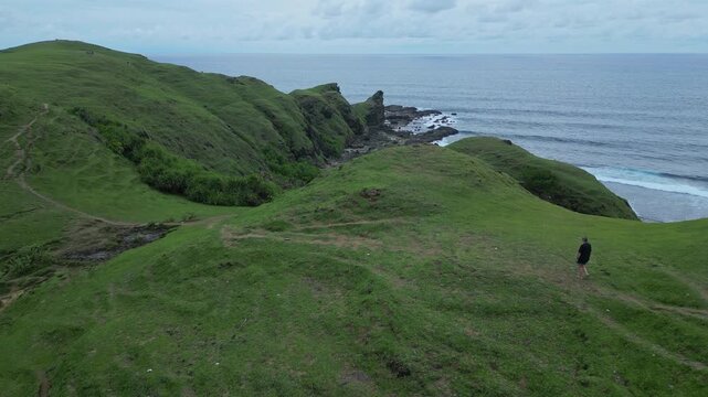 Woman Walking Cliffside Path Toward Headland, Rocky Promontory And Open Sea Frame Steady Forward Stride, Expansive Coastal Vista, Travel And Adventure Tone With Rugged Green, Lombok