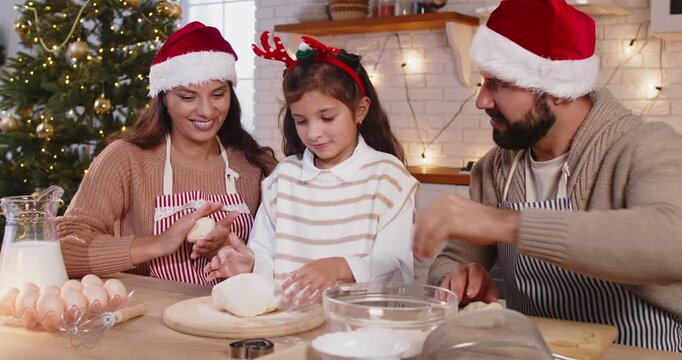 Happy family baking Christmas cookies together for winter season at home kitchen. Parents in aprons and Santa hats help child to knead dough and sift flour, bake pastry according to traditional recipe